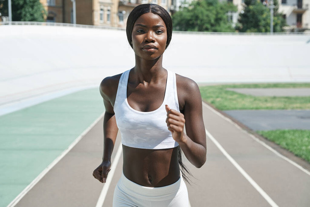 Beautiful young African woman in sports clothing running on track outdoors - Fotó, kép
