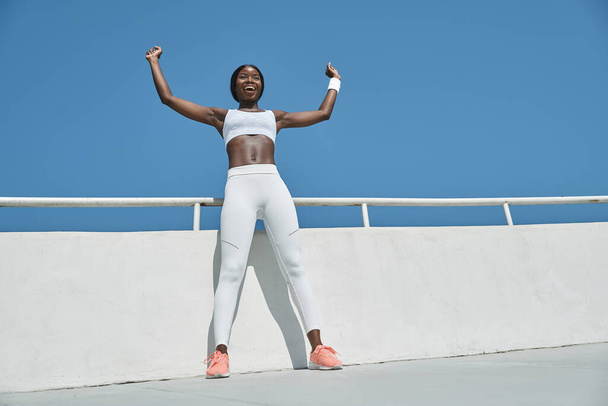Low angle view of beautiful young African woman in sports clothing keeping arms raised and smiling - Photo, Image