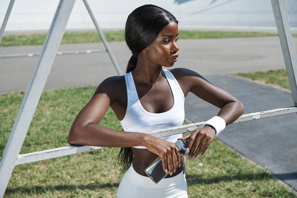 Beautiful young African woman in sports clothing holding bottle with water while standing outdoors - Φωτογραφία, εικόνα