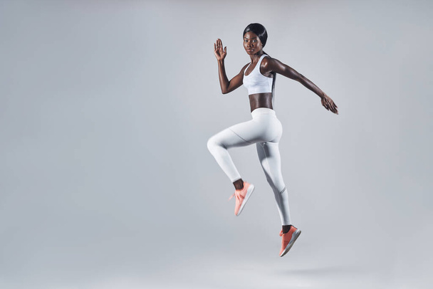 Full length of confident young African woman in sports clothing jumping against gray background - Foto, Imagen