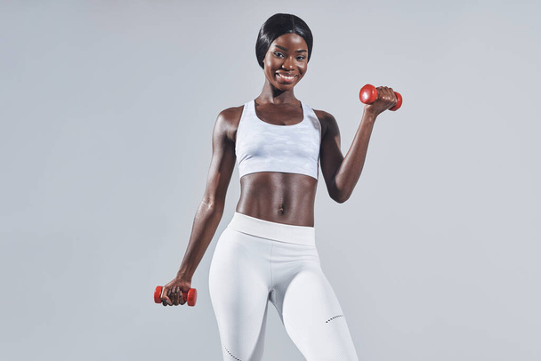 Beautiful young African woman in sports clothing exercising with dumbbells - 写真・画像