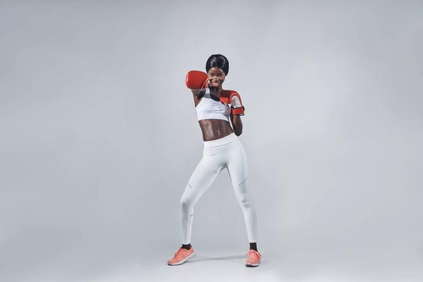 Beautiful young African woman in boxing gloves exercising while standing against gray background - Φωτογραφία, εικόνα