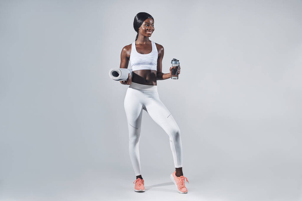 Happy young African woman in sports clothing holding bottle with water and exercise mat - Foto, Imagem