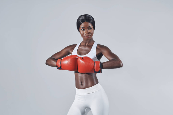 Beautiful young African woman in boxing gloves standing against gray background - 写真・画像