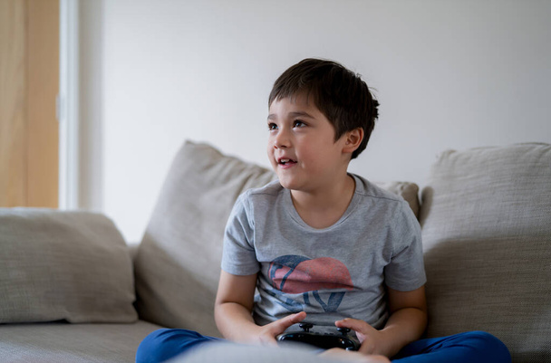 若い男の子は、オンラインでビデオゲームをプレイし、カンディードはソファを保持するゲームコンソールに座ってハッピー混合レースの子供を撮影しました。週末に家でリラックスしている間に肖像画の子供の顔 - 写真・画像