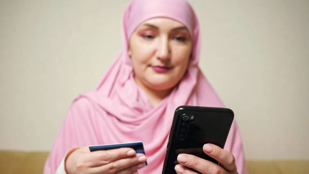 Γυναίκα σε ένα ροζ κασκόλ κάνει online αγορές στο τηλέφωνο με μια τραπεζική κάρτα στα χέρια - Φωτογραφία, εικόνα