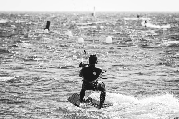 Genç Akdeniz erkeği uçurtma uçuruyor. Siyah bir dalış kıyafetiyle suda ve uçurtmayı uçurmak üzere. Uçurtmanın siyah ve beyaz çekimi. - Fotoğraf, Görsel