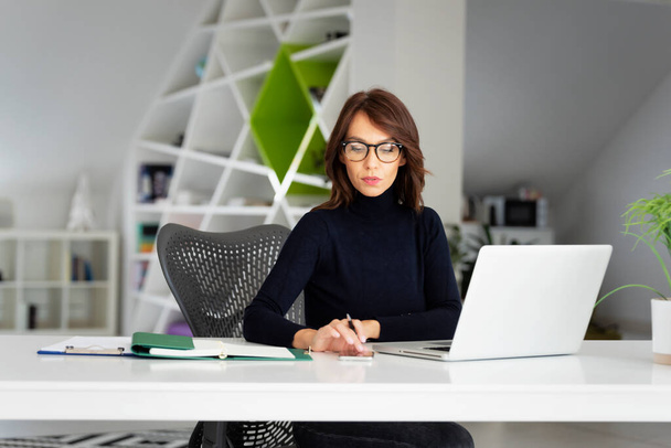 Confident middle aged businesswoman wearing turtleneck sweater while sitting at office desk behind her laptop and working on new project.  - 写真・画像