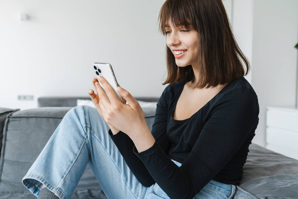 Smiling young woman using mobile phone while sitting on a couch at home - Фото, изображение