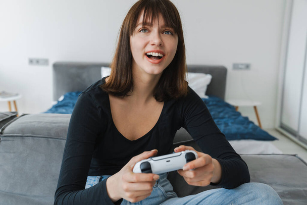 Happy young white woman holding joystick sitting on a couch at home - Zdjęcie, obraz