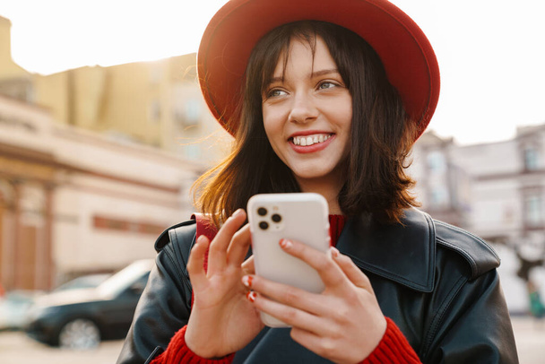 Happy young brunette white woman standing on a street, holding mobile phone - Foto, Bild