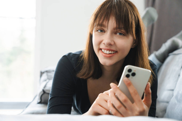 Smiling young woman holding mobile phone texting while laying on couch at home, close up - Photo, image