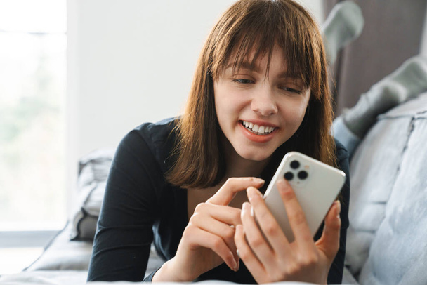 Smiling young woman using mobile phone while laying on a couch at home - Photo, Image