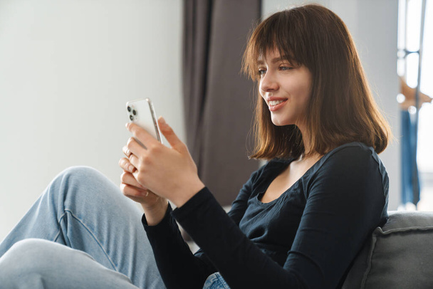 Smiling young woman holding mobile phone texting while sitting leaning on a couch at home - Foto, immagini