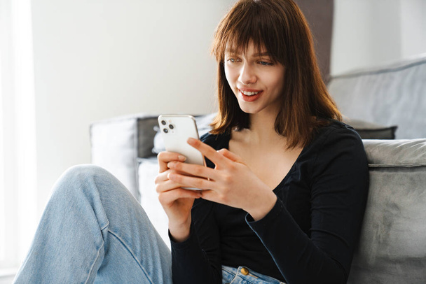 Smiling young woman holding mobile phone texting while sitting leaning on a couch at home - Photo, image