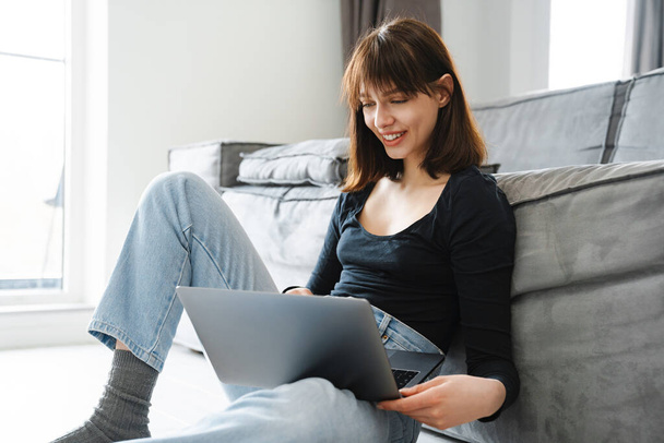 Smiling young white woman with a laptop sitting on a floor at home - Photo, Image