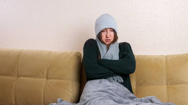 Woman with plaid puts on scarf sitting on sofa in cold room - Photo, Image