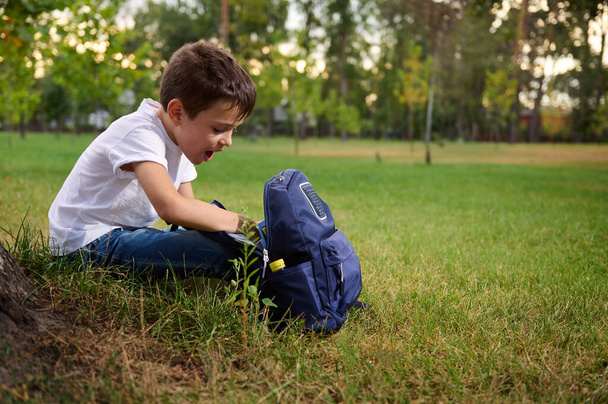 Elementary aged school boy in casual denim resting on green grass after his first day at school. Adorable child with backpack during recreation on beautiful sunny day. Back to school concept - Photo, Image