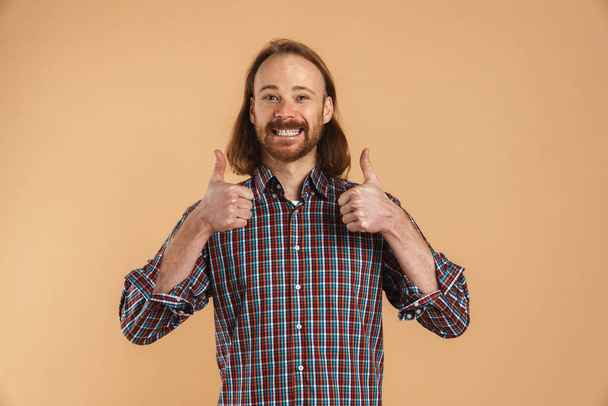 Bearded ginger man wearing plaid shirt smiling and showing thumbs up isolated over beige background - Foto, Bild