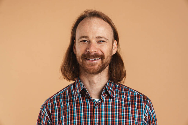 Рыжий с бородой мужчина в клетчатой рубашке улыбается и смотрит на камеру, изолированную на бежевом фоне - Фото, изображение