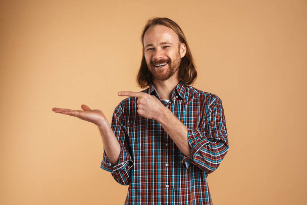 Smiling ginger man holding and pointing finger at copyspace isolated over beige background - Foto, Bild