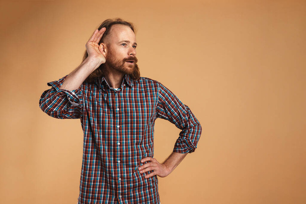 Bearded ginger focused man gesturing and eavesdropping isolated over beige background - Foto, immagini