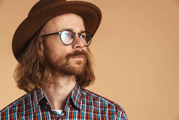 Bearded ginger man wearing eyeglasses and hat looking aside isolated over beige background - Foto, Bild