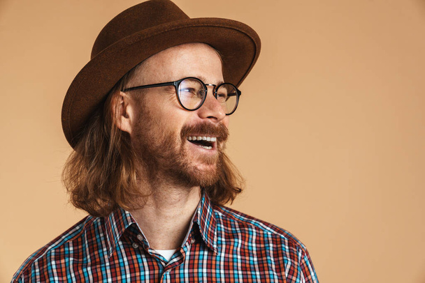 Laughing ginger man wearing eyeglasses and hat looking aside isolated over beige background - Fotografie, Obrázek