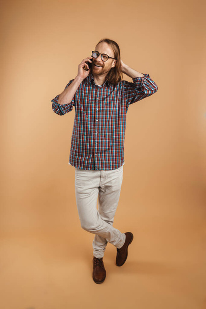 White ginger man wearing eyeglasses smiling while talking on cellphone isolated over beige background - Photo, image