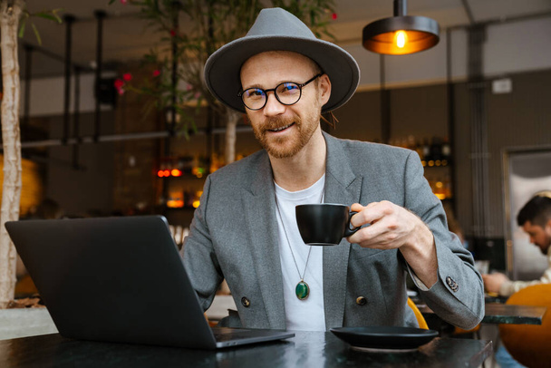 White bearded man in jacket drinking coffee and using laptop in cafe outdoors - Foto, immagini
