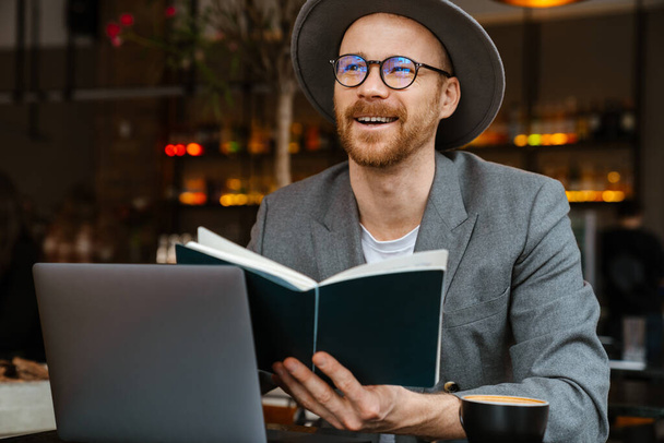 Smiling mid aged fashionable hipster man wearing glasses and hat sitting at the cafe table, reading book - Foto, Bild