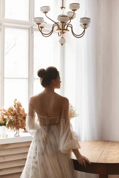 Молодая модель женщина в винтажном свадебном платье с обнаженной спиной позирует в винтажном интерьере в помещении. Стильная молодая невеста в модном винтажном платье позирует со спиной - Фото, изображение