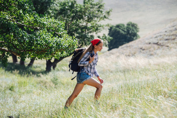 Uphill walk of a woman with a read headband hiking in grassy hills. Side view. She's wearing small backpack. - Zdjęcie, obraz