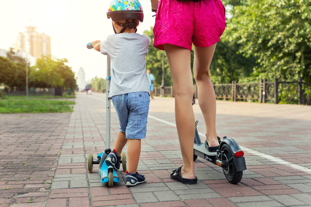 Vista trasera trasera de lindo niño pequeño con madre joven adulta montando transporte de scooter en el parque de la calle de la ciudad juntos en el día soleado. Atención y apoyo a los padres. Concepto de estilo de vida saludable para niños activos - Foto, imagen