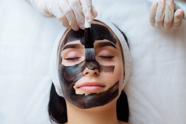 косметолог надевает черную маску на красивую женщину в черных перчатках, великолепная женщина в спа проходит процедуры по уходу за лицом
. - Фото, изображение