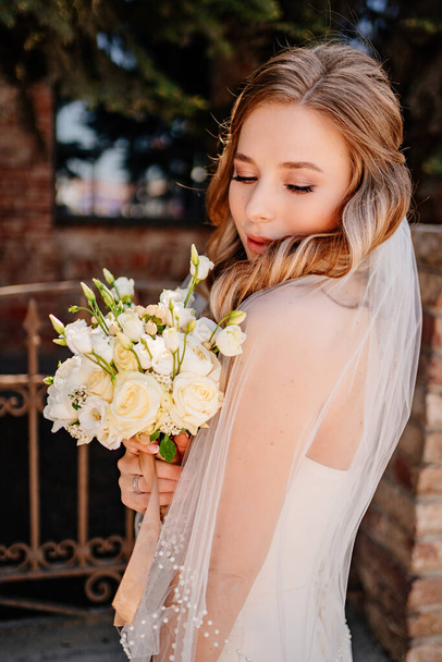 gentle and beautiful bride in a veil with a bouquet of flowers at the fence. - Foto, Bild