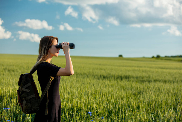 mladá dívka v černých šatech s dalekohledem a batohem na zeleném pšeničném poli - Fotografie, Obrázek