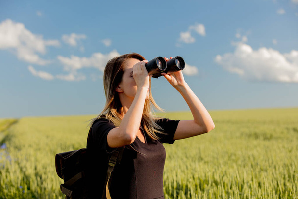 young girl in black dress with binoculars and a backpack on a green wheat field - Foto, Bild