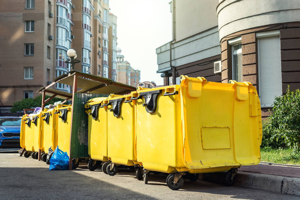 Reihenweise große gelbe Plastikmülltonnen voller schwarzer Plastik-Müllsäcke in der Nähe von Wohnhäusern in der Innenstadt oder am Stadtrand. Nicht recycelbare Mülltrennung - Foto, Bild