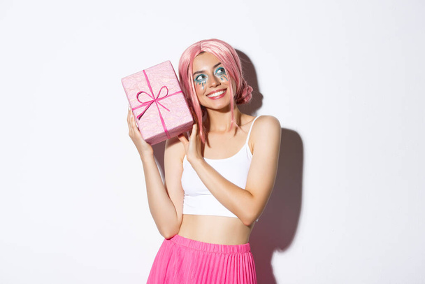 Immagine di bella ragazza in parrucca rosa scuotendo scatola con regalo di compleanno, chiedo cosa all'interno scatola avvolta, in piedi su sfondo bianco - Foto, immagini
