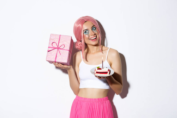 Imagem da menina bonito animado em peruca rosa, agitando caixa com presente e passear o que dentro, segurando pedaço de bolo de aniversário, celebrando b-dia - Foto, Imagem