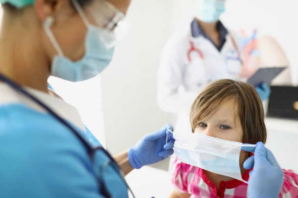 vrouw dokter in een masker zet een masker op een kind - Foto, afbeelding