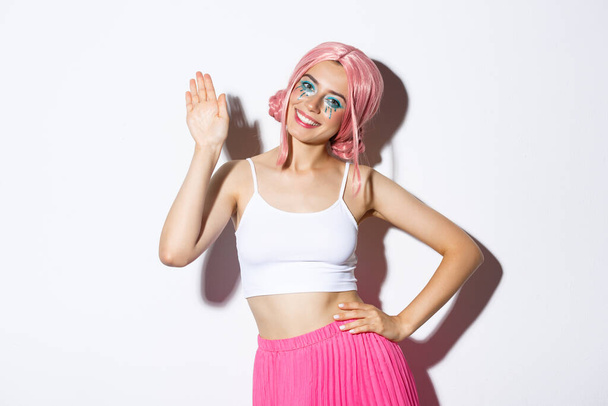 Imagen de la alegre chica glamour con peluca rosa y maquillaje de Halloween, saludando, saludando con la mano y saludando a alguien, de pie sobre fondo blanco - Foto, Imagen