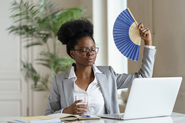 Exhausted businesswoman tired of heat waving paper fan for fresh air at workplace work on laptop - Photo, image