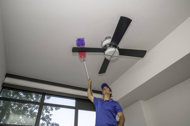 Homme nettoie les lames de ventilateur de plafond poussiéreux à l'intérieur d'une maison - Photo, image