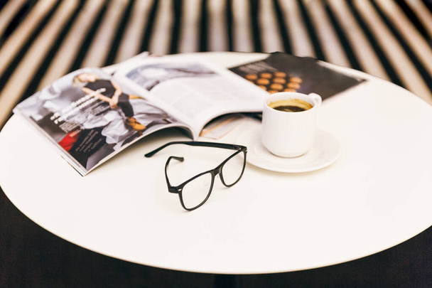 Обрезанные фото белого стола с некоторыми журналами, чашка кофе и избирательный акцент на очки / заряд энергии утром, вкусный горячий черный кофе за столом, на рабочем месте, кофе кружку. - Фото, изображение