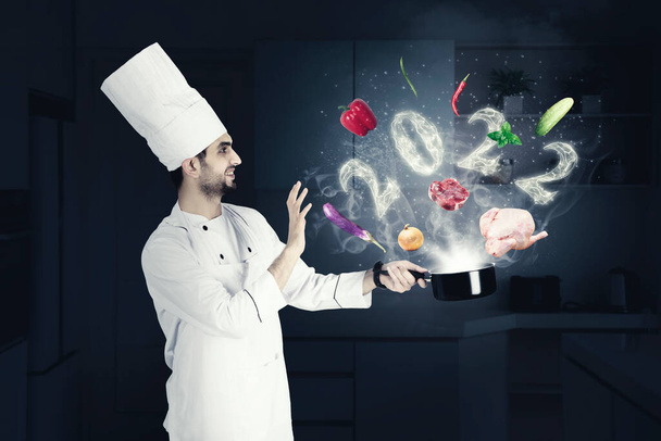 Happy male magician chef cooking on a saucepan with flying food ingredients and smoke shaped of 2022 numbers in the kitchen - 写真・画像