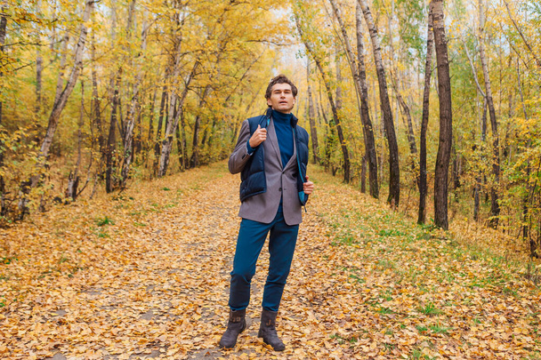 Tall handsome man dressed in a brown jacket walking in the autumn alley - Foto, afbeelding