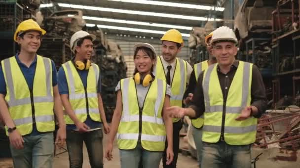 Various races workers and engineers of factory are having fun joking around in machine-spare parts warehouse during break. The entire team wears safety uniforms and industrial helmets. - Footage, Video