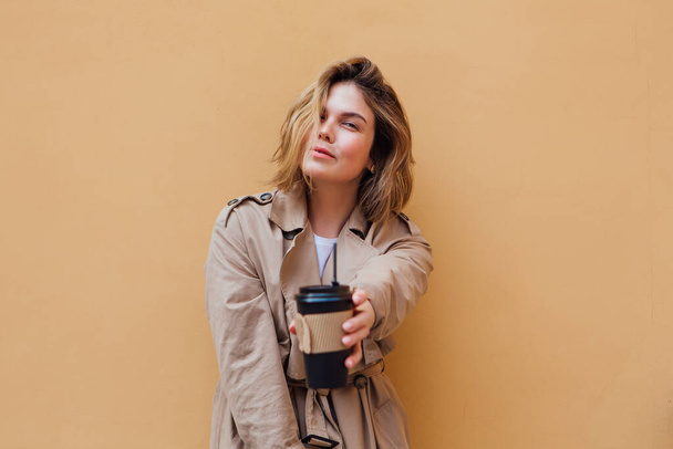 Young smiling millennial woman with wild hair dressed in an autumn coat standing a cup of coffee to go near the beige wall. - Foto, Imagen